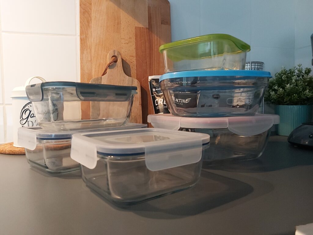 6 récipients hermétiques en verre avec couvercle pour le meal prep, posés dans une cuisine avec en arrière-plan des planches à découper, un contenant pour les gousses d'ail et un sachet de créatine